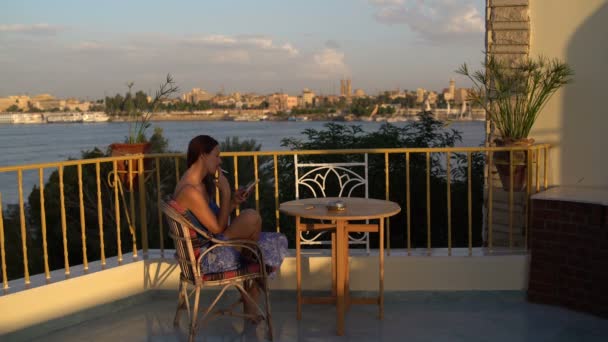 Vrouw zit op het terras met uitzicht op de rivier rookt en maakt gebruik van de smartphone — Stockvideo