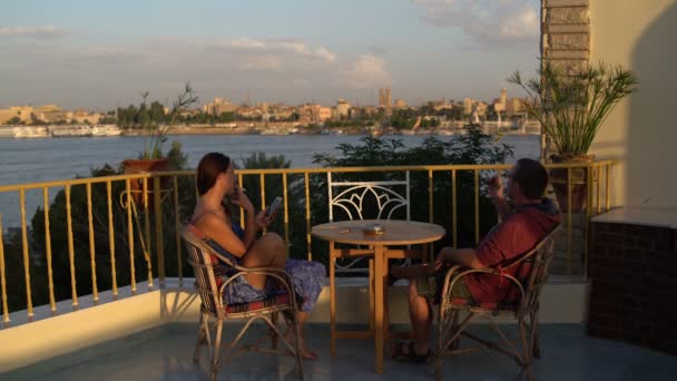 Мужчина и женщина сидят за столом на террасе с видом на реку. Женщина курит — стоковое видео