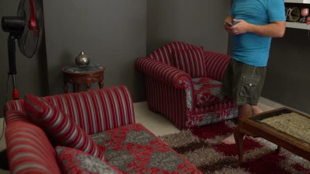 Un hombre enciende un ventilador y se sienta en una silla con un teléfono inteligente — Vídeo de stock