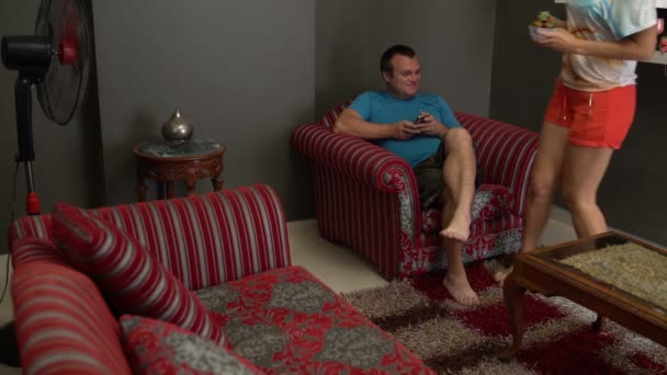 Een man zit in een stoel met een smartphone. Een vrouw wordt geleverd met een aardbei en behandelt een man — Stockvideo