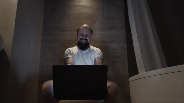 En man skrattar sittandes vid en dator på toaletten — Stockvideo