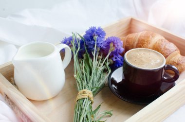 Romantik yaz odaya yatak, taze kruvasan ile tepsi, fincan sütlü kahve espresso ve mavi Peygamber buket. Günaydın kavramı. Yavaş hayattan zevk. Geleneksel kahvaltı.