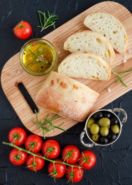 Biberiye, zeytin yağı, domates ile ahşap kesme tahtası üzerinde taze pişmiş ciabatta ekmeği dilimlenmiş, Siyah Zeytin ve tuz karanlık taş beton tablo. İtalyan gıda kavramı. Üstten Görünüm.