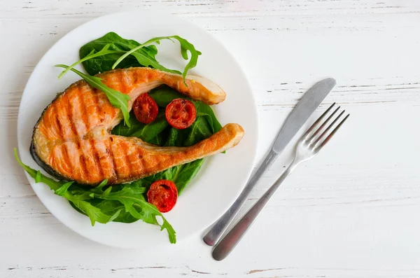 Ispanak, kiraz domates ve bıçak ile bir tabak hizmet üzerinde roka ve çatal Beyaz ahşap zemin üzerinde ızgara somon steak. Sağlıklı öğle yemeği kavramı. Yemek lezzetli, besleyici. Üstten Görünüm.