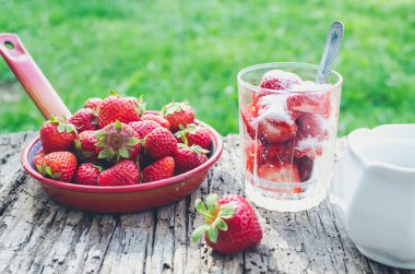 Bir kaşık ile bir bardak çilek ile şeker yemeye hazır. Sulu sulu taze Olgun kırmızı çilek bir eski ahşap dokulu masa üstü arka planını, yeşil çim üzerinde. Dışarıda kahvaltı.