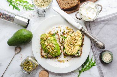 Kahvaltıda sağlıklı avokado tostları ya da öğle yemeğinde çavdar ekmeği, krem peynir, roka, dilimlenmiş avokado, balkabağı, kenevir ve susam tohumu, tuz ve biber. Vejetaryen gıda konsepti. Temiz yemek. Üst görünüm.