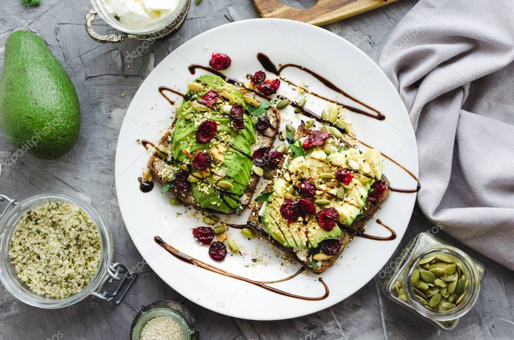 Healthy avocado toasts for breakfast or lunch with rye bread, cream cheese, arugula, sliced avocado, dried cranberry, pumpkin, hemp and sesame seeds. Vegetarian sandwiches. Clean eating. Top view.