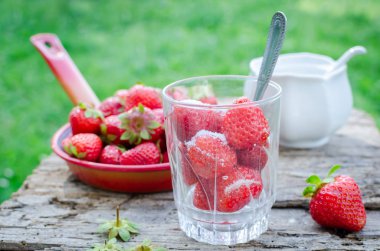 Bir kaşık ile bir bardak çilek ile şeker yemeye hazır. Sulu sulu taze Olgun kırmızı çilek bir eski ahşap dokulu masa üstü arka planını, yeşil çim üzerinde. Dışarıda kahvaltı.