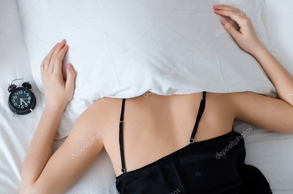Young woman with pillow over her head with alarm clock on bed in the morning. Student or schoolgirl do not want to wake up early for school or univercity. Oversleep, not getting enough sleep concept.