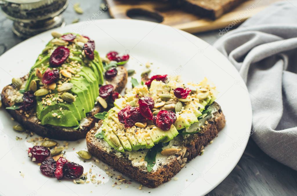 Healthy avocado toasts for breakfast or lunch with rye bread, cream cheese, arugula, sliced avocado, dried cranberry, pumpkin, hemp and sesame seeds. Vegetarian sandwiches. Clean eating concept.