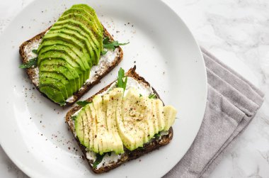 Dilimlenmiş avokado, roka, tuz ve biber hafif mermer zemin üzerine sağlıklı kahvaltı ile krem peynir ile siyah ekmek üzerinde avokado tost. Vejetaryen yiyecekler. Bitki temelli diyet. Temiz yemek. Üstten görünüm.