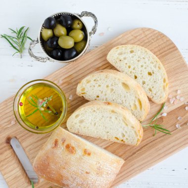 Biberiye, tuz, zeytin ve zeytinyağı ile beyaz rustik tabloda ahşap kesme tahtası üzerinde taze pişmiş ciabatta ekmeği dilimlenmiş. İtalyan gıda kavramı. Üstten Görünüm.