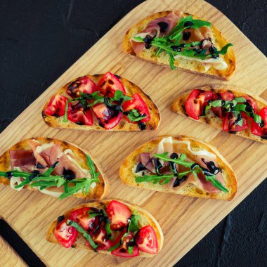 Lezzetli İtalyan bruschetta veya kanepeler üzerinde tost baget Prosciutto di Parma, roka ve balzamik glasse üzerinde kesme tahtası koyu taş zemin üzerine sos domates ile tepesinde seçimi. Üstten Görünüm.