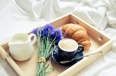 Romantik yaz odaya yatak, taze kruvasan ile tepsi, fincan sütlü kahve espresso ve mavi Peygamber buket. Günaydın kavramı. Yavaş hayattan zevk. Geleneksel kahvaltı.