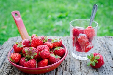 Bir kaşık ile bir bardak çilek ile şeker yemeye hazır. Sulu sulu taze Olgun kırmızı çilek bir eski ahşap dokulu masa üstü arka planını, yeşil çim üzerinde. Dışarıda kahvaltı.