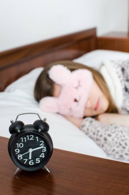 Genç kadın kız sevimli uyku maskesi ve sıcak pijama uyku ile çalar saat sabah yatakta yakınındaki. Uyuyan kız ve alarmclock yatak odasında. Genç geç uyanmak korkuyor. Kavram fazla uyumak.