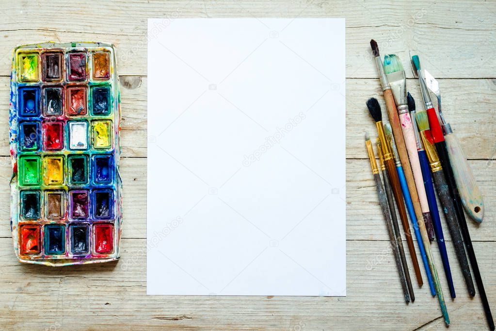 Artist paint brushes and watercolor paintbox on wooden background with empty sheet of paper. Instruments and tools for creative leisure. Paintings art concept mockup. Top view. Copy space. Flat lay.