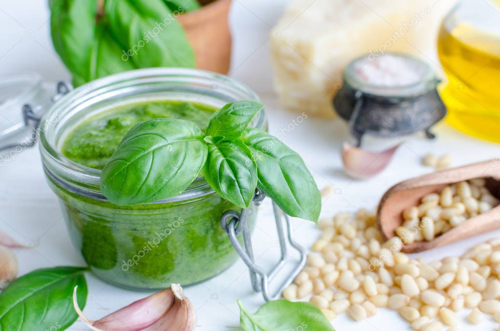 Pesto genovese - traditional Italian green basil sauce with raw ingredients on white wooden background. Basil leaves in mortar, Parmesan cheese, pine nuts, olive oil, garlic and salt.