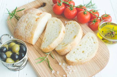 Biberiye, tuz, zeytin, domates ve zeytinyağı ile beyaz rustik tabloda ahşap kesme tahtası üzerinde taze pişmiş ciabatta ekmeği dilimlenmiş. İtalyan gıda kavramı.