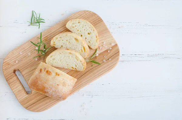 Biberiye ve tuz ile beyaz rustik tabloda ahşap kesme tahtası üzerinde taze pişmiş ciabatta ekmeği dilimlenmiş. Üstten görünüm. Kopya alanı.