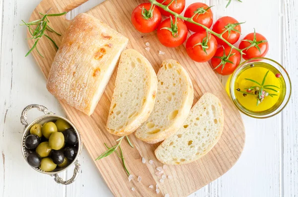 Biberiye, tuz, zeytin, domates ve zeytinyağı ile beyaz rustik tabloda ahşap kesme tahtası üzerinde taze pişmiş ciabatta ekmeği dilimlenmiş. İtalyan gıda kavramı. Üstten görünüm.