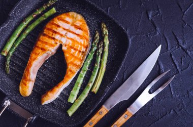 Izgara somon steak ve grill Pan siyah taş zemin üzerine bıçak ve çatal ile hazırlanmış kuşkonmaz. Sağlıklı gıda kavramı. Üstten görünüm.
