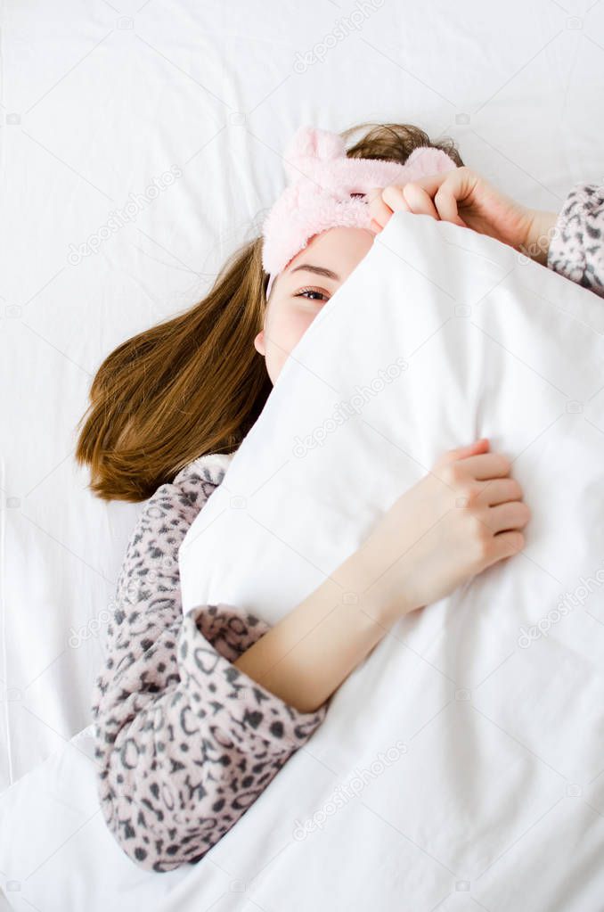 Happy young woman enjoying sunny morning hugging pillow. Rest, sleeping, comfort concept - caucasian girl lying in the bed at home bedroom and smiling after wake up. View from above.