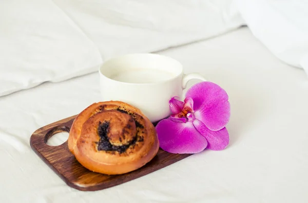 Cup of coffee cappuccino with pastry on wooden board on a bed on cozy lazy sunday. White bedding sheet, blanket and pillows. Good morning concept. Enjoy slow life.