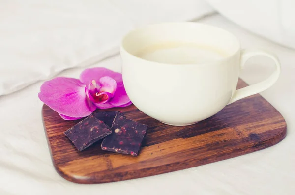 Cup of coffee cappuccino with chocolate and flower on wooden board on a bed on cozy lazy sunday. White bedding sheet, blanket and pillows. Good morning concept. Enjoy slow life.