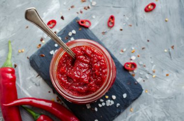 Kırmızı sıcak biber reçeli cam gri beton zemin üzerine taze malzemelerle. Doğal ev yapımı biber sosu kaşıkla. Taze ev yapımı salsa sosu. Üstten görünüm.