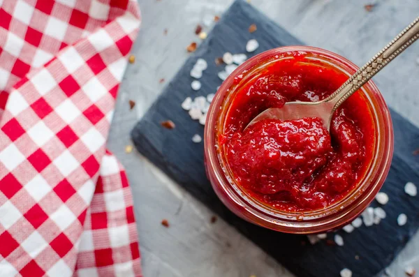 Kırmızı sıcak biber reçeli cam gri beton zemin üzerine taze malzemelerle. Doğal ev yapımı biber sosu kaşıkla. Taze ev yapımı salsa sosu. Üstten görünüm. Yakın çekim.