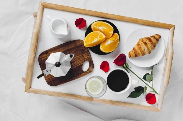 Romantic Valentines Day breakfast in bed, tray with croissant, cup of coffee espresso with milk, fresh oranges, glass of water with lemon and red rose. Good morning concept. Enjoy slow life. Top view.