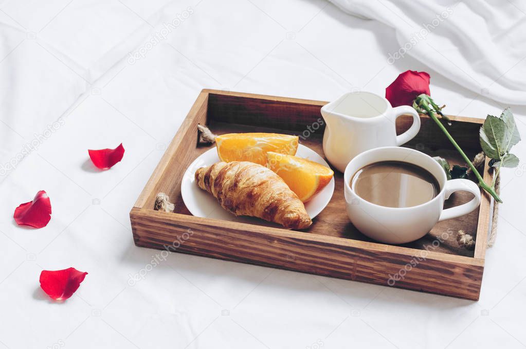 Romantic Valentines Day breakfast in bed, tray with fresh croissant, cup of coffee espresso with milk, fresh oranges and red rose. Good morning concept. Enjoy slow life.