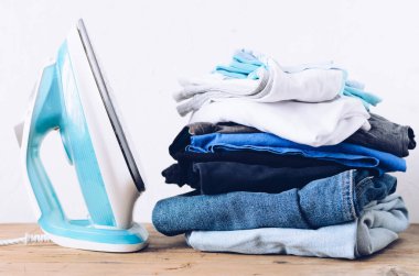 Kapalı masada giysi ve elektrikli demir yığını. Ev konsepti. Taze katlanmış pamuk kot giyim. Ütü, giysi, ev işi konsepti - evde masada demir ve giysi yakın.