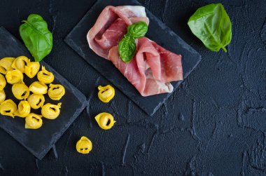 Taze hazırlanmış çiğ ev yapımı et dolu prosciutto tortellini fesleğen yaprakları siyah arka plan üzerinde koyu taş tabakta yemek hazır. Geleneksel İtalyan mutfağı kavramı. Üstten Görünüm. Kopya alanı.