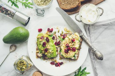 Kahvaltıda sağlıklı avokado tostları ya da öğle yemeğinde çavdar ekmeği, krem peynir, roka, dilimlenmiş avokado, kızılcık, balkabağı, kenevir ve susam tohumu. Vejetaryen sandviçler. Temiz yemek. Üst görünüm.