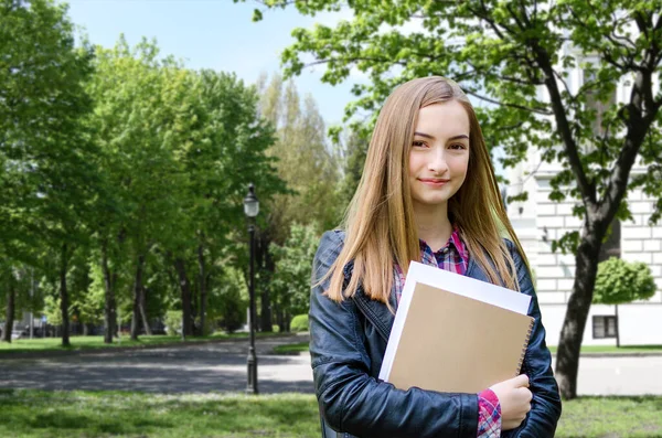 Young girl high school or college student holding textbooks, smiling and looking at the camera. She is on her way to class near university building. Back to school concept.