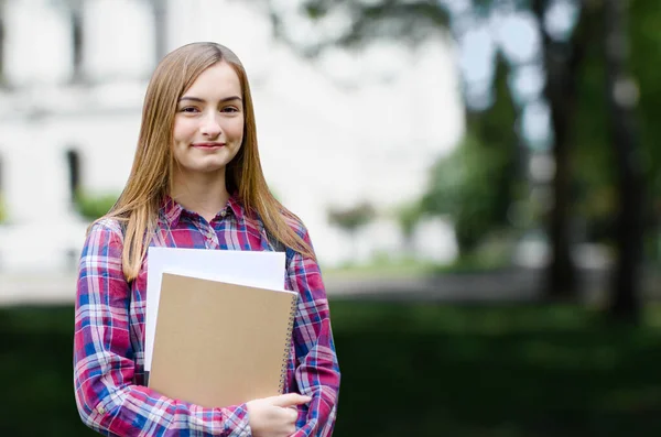 Young girl high school or college student holding textbooks, smiling and looking at the camera. She is on her way to class near university building. Back to school concept. Blurred background.