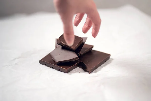 Hand grabbing a chocolate piece, cut chocolate on a white paper background, having a dark chocolate as a snack