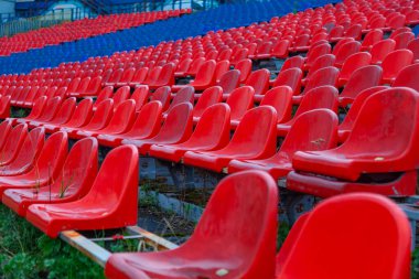 Plastik kırmızı ve mavi koltukları olan eski bir futbol stadyumunun tribünü. Sandalyelerin bir kısmı kırılmış.