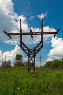 A rusty old electric transmission line pylon stands in a field against a cloudy sky. In the background is a modern electric transmission line support