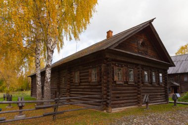 Old log house, blackened from time to time, surrounded by a wooden fence. Near the house there is a birch with yellow leaves.