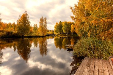 Autumn landscape - trees with yellowed leaves stand on the shores of the lake in which their reflection is visible. In the foreground is a wooden bridge