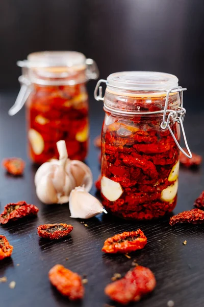 Sun-dried tomatoes are packed in a jar with spices and garlic and drenched in olive oil. Tins of tomatoes are on a black table, with slices of tomato and spices scattered nearby.