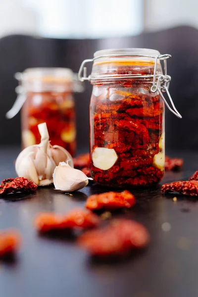 Sun-dried tomatoes are packed in a jar with spices and garlic and drenched in olive oil. Tins of tomatoes are on a black table, with slices of tomato and spices scattered nearby.