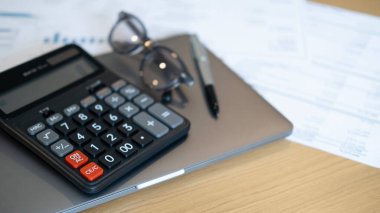 A black calculator with glasses and a pen on a gray laptop surrounded by financial reports symbolizing careful management accounting and preparation for long term security.