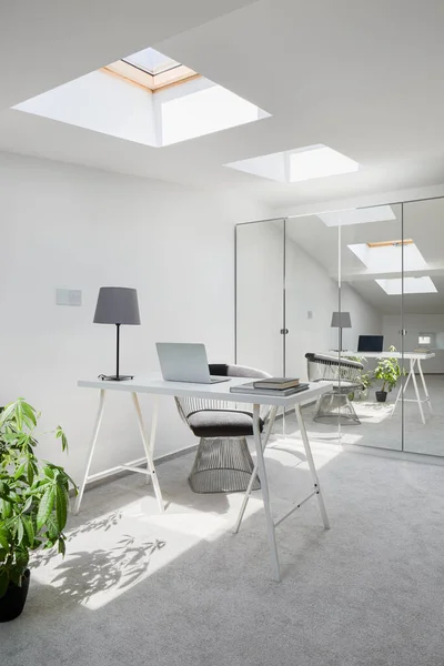 Office area with simple white desk in attic room with ceiling windows, gray flooring and wardrobe with mirrored doors
