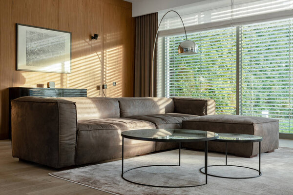 Elegant living room with big corner sofa, two modern coffee tables and silver lamp with big windows with garden view