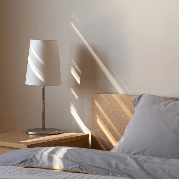 Simple, white lamp on wooden bedside table next to bed with wooden headboard and gray bedclothes