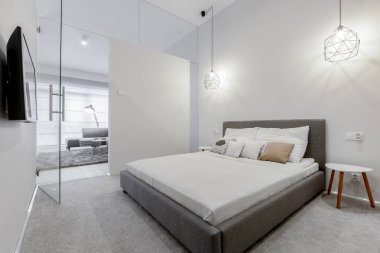 Parlak beyaz yatak odasında gri çerçeveli büyük bir yatak ve oturma odasına açılan cam kapılar.